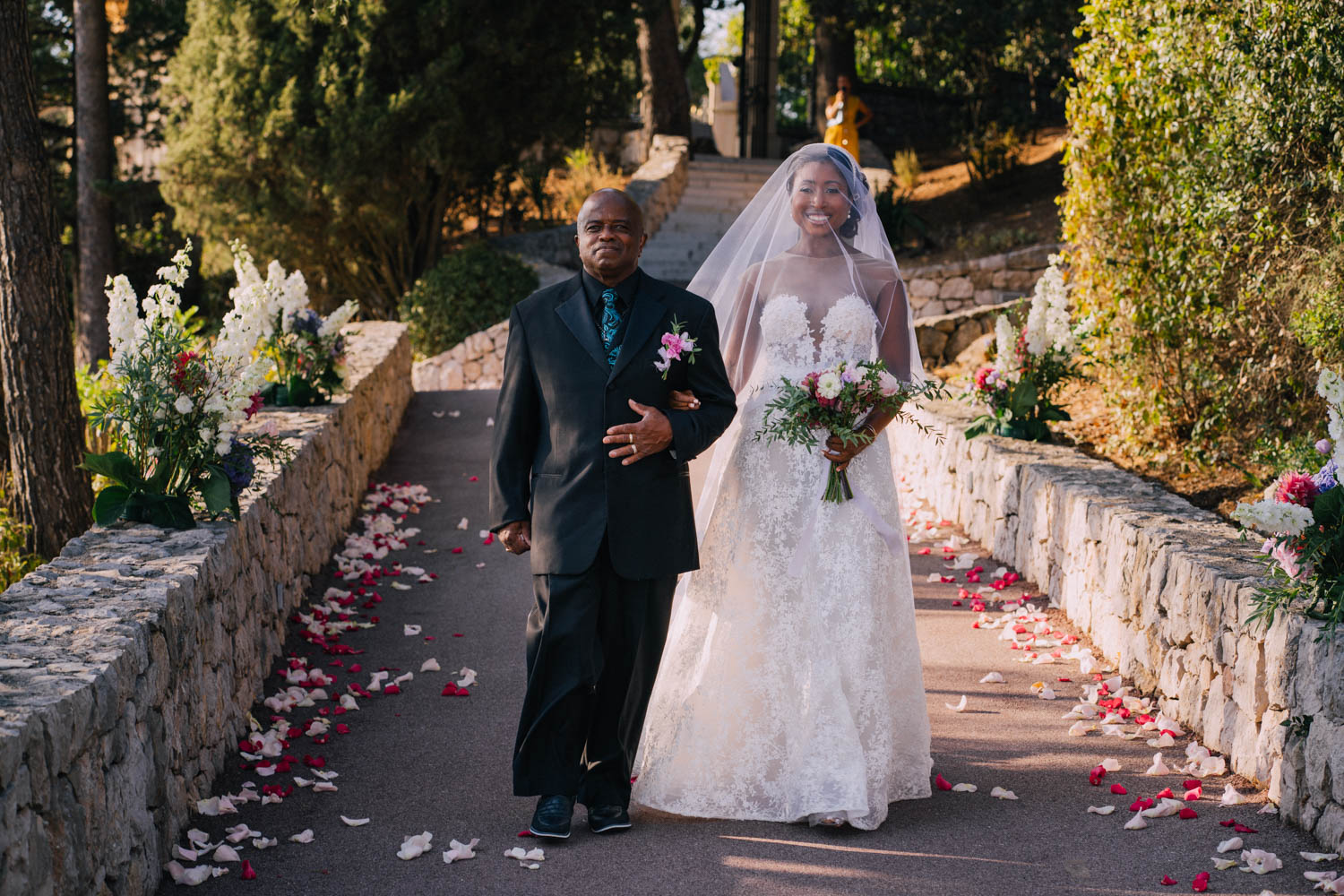 Dad in a black suit walks his happy bride down the aisle lined with rose petals