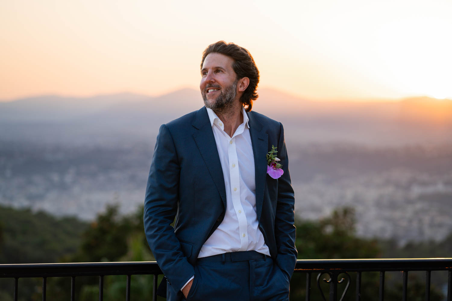 A groom leans on a balcony railing in a blue suit smiling at sunset