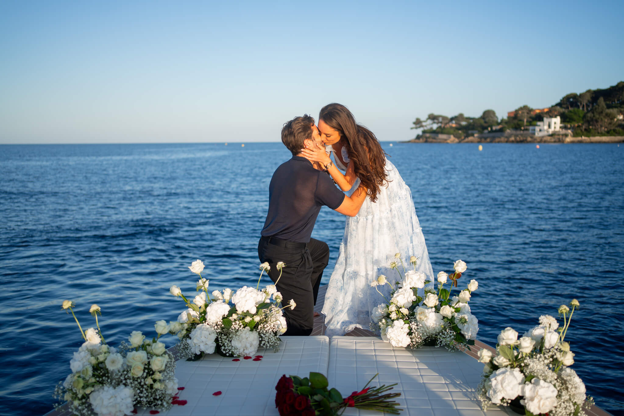 Newly engaged couple kisses while on one knee on the bow of a boat