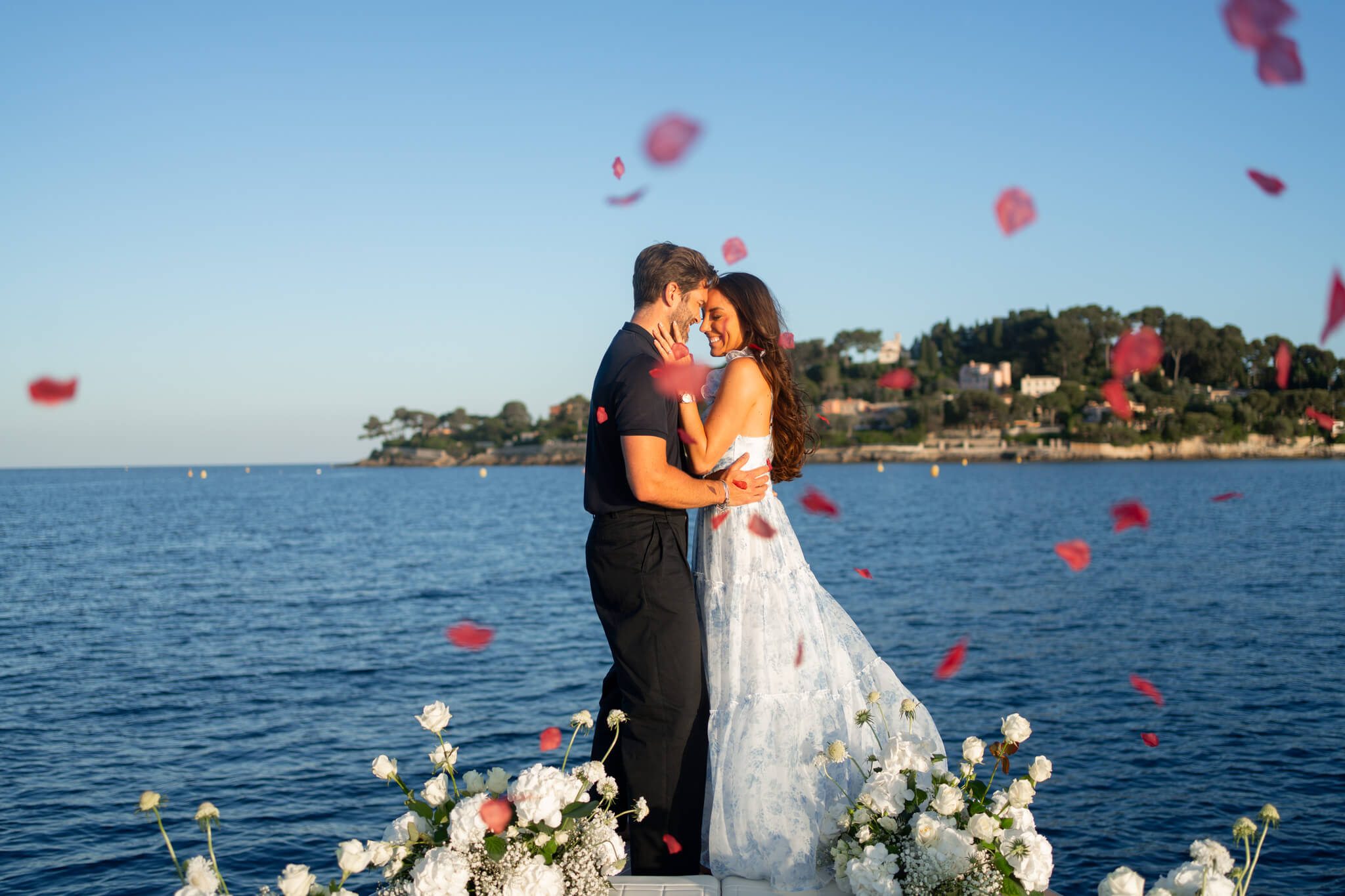 A happy couple snuggles after their French Riviera Proposal on the bow of a boat as red rose petals flie around them