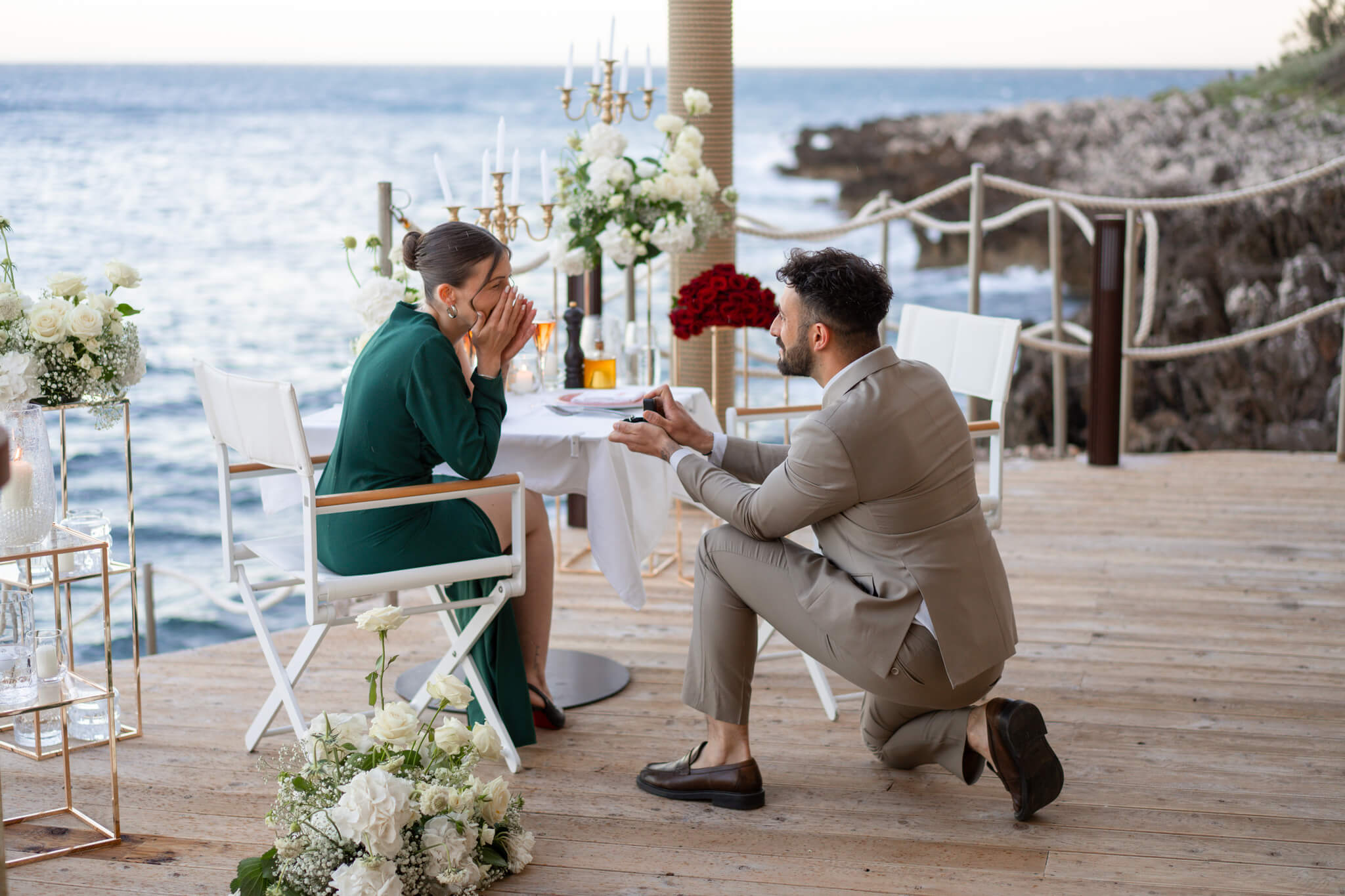 A man in a tan suit gets down on one knee to present his fiancee a ring as she covers her face in shock