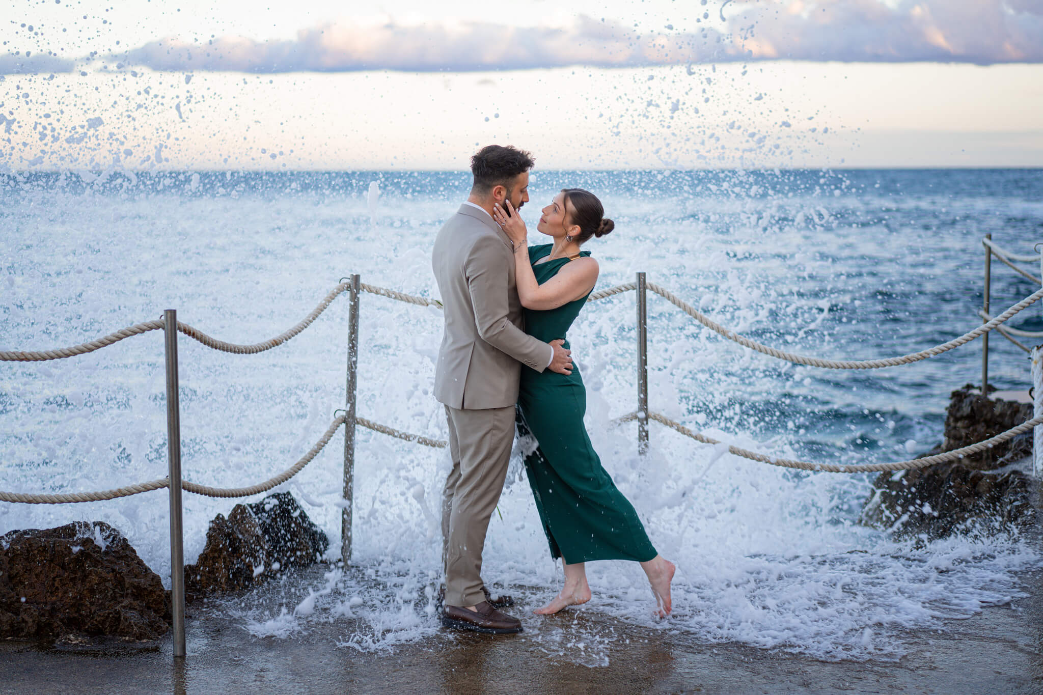 A happy couple embrace as waves crash around them during their French Riviera Proposal